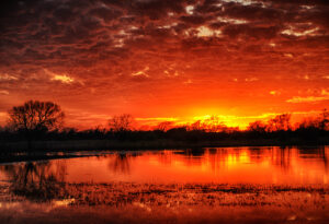 A vibrant sunset with deep reds and oranges reflecting off a calm lake, by Ken Osborne.