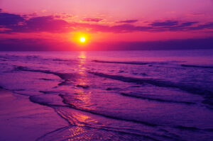 Vibrant sunset over calm seas with waves gently approaching the shore under a dramatic purple sky.