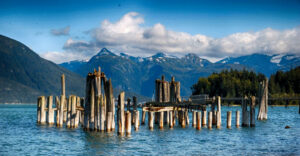 Weathered wooden pillars of an old dock stand in Alaskan waters with mountains in the distance