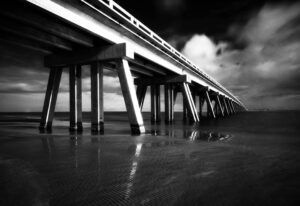 Monochrome image of San Luis Bridge extending into the distance over a smooth beach.