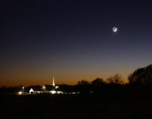 The crescent moon hanging over a lit-up church in the stillness of Simonton night