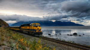 A train traveling alongside a serene Alaskan lake with mountains in the backdrop in Ken Osborne's photograph