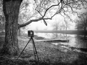 A vintage large format camera poised on a tripod by the misty banks of Bessie's Creek, in a monochrome image by Ken Osborne.