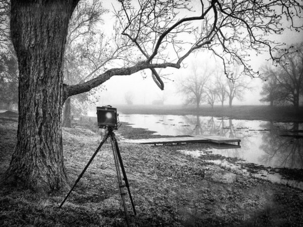 A vintage large format camera poised on a tripod by the misty banks of Bessie's Creek, in a monochrome image by Ken Osborne.